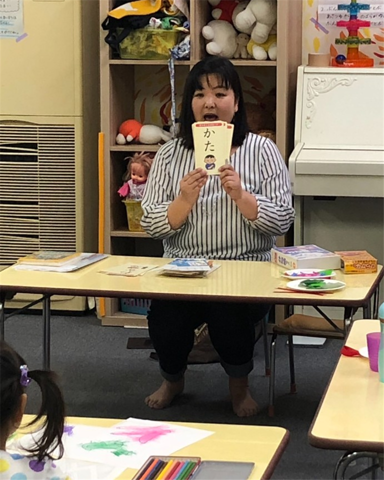 支援員が絵カードで手遊びをリードし、お子さまの後ろ姿から参加するワークショップの様子